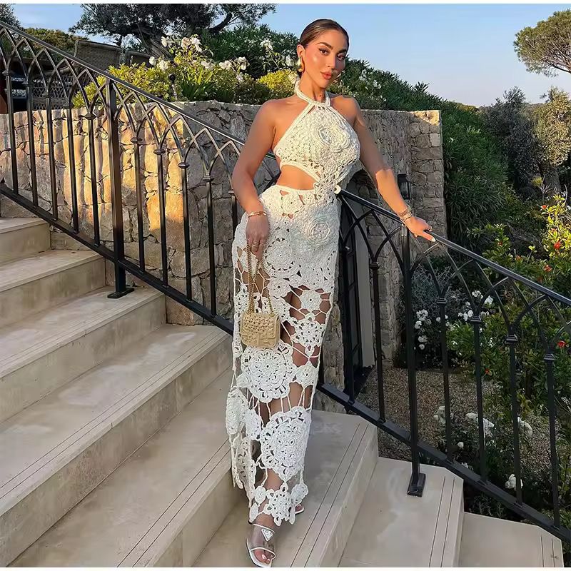 Woman standing on outdoor stone stairs wearing a white floral crochet halter dress with cut-outs and open back. The ankle-length dress is semi-sheer, styled with white heels and a small woven handbag. Surrounded by lush greenery and Mediterranean stone architecture in golden hour lighting.