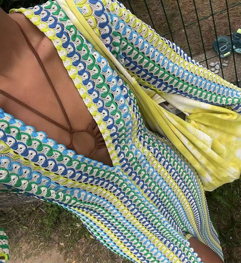 Woman wearing a colorful crochet knit maxi dress with vertical rainbow stripes, deep V-neckline, flared long sleeves, and high leg slit – standing confidently on a sunny beach, styled for summer vacations, resort wear, boho chic fashion, and Italian coastal vibes.