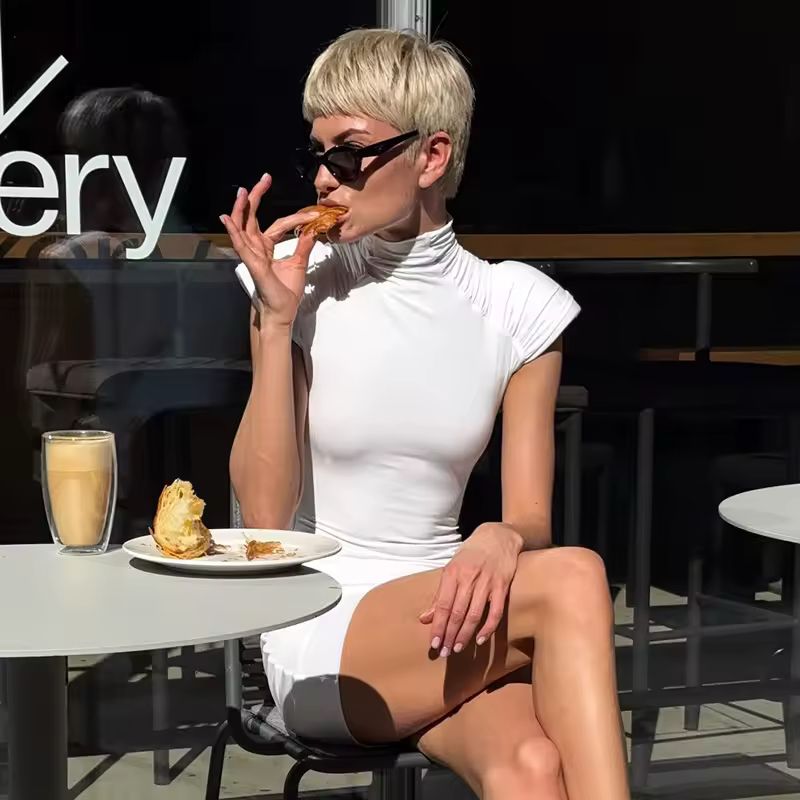 Woman wearing white high-neck sleeveless mini dress with ruched shoulders, sitting in sunlight.