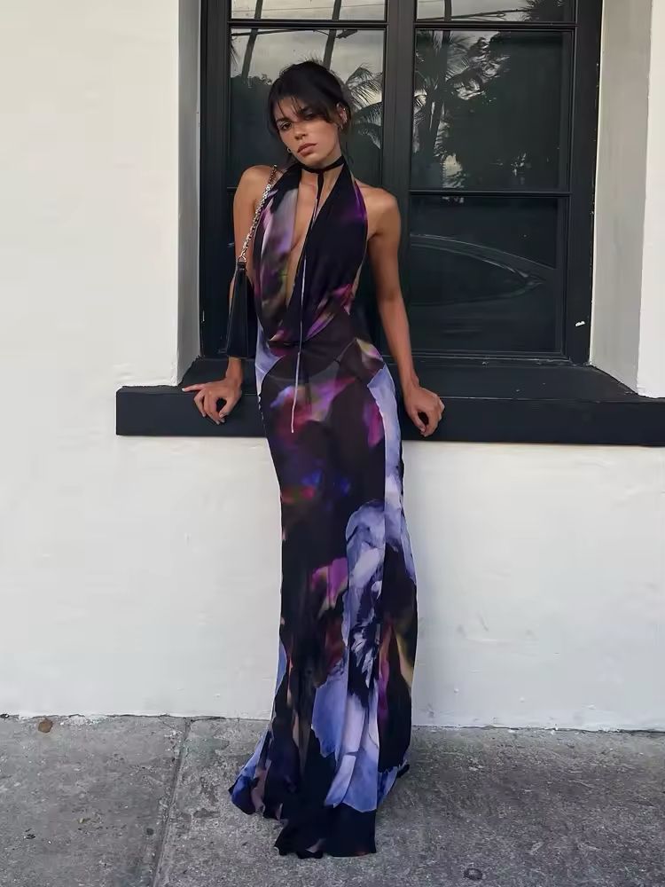Collage of four images showcasing the “Midnight Bloom” dress. The model wears a backless, deep V-neck floral maxi dress in dark tones with purple, blue, and pink abstract flower prints. Close-up shots highlight the flowy fabric and elegant silhouette. The setting includes minimalist architecture and seaside views.