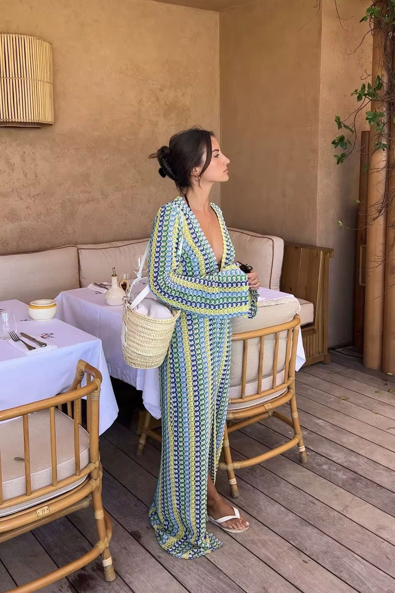 Woman wearing a colorful crochet knit maxi dress with vertical rainbow stripes, deep V-neckline, flared long sleeves, and high leg slit – standing confidently on a sunny beach, styled for summer vacations, resort wear, boho chic fashion, and Italian coastal vibes.