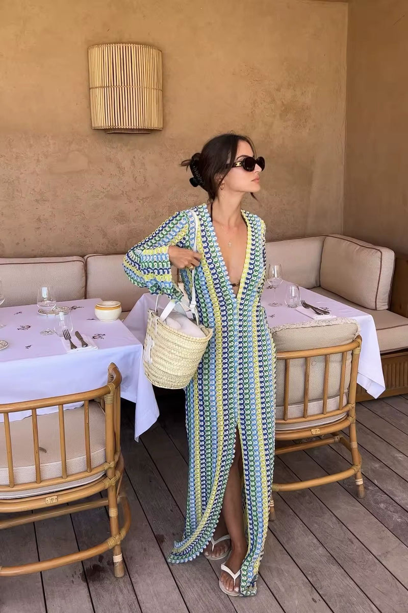 Woman wearing a colorful crochet knit maxi dress with vertical rainbow stripes, deep V-neckline, flared long sleeves, and high leg slit – standing confidently on a sunny beach, styled for summer vacations, resort wear, boho chic fashion, and Italian coastal vibes.