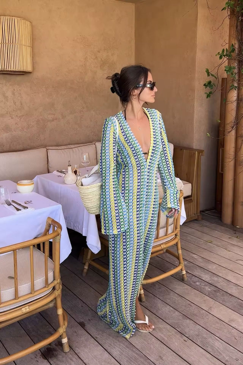 Woman wearing a colorful crochet knit maxi dress with vertical rainbow stripes, deep V-neckline, flared long sleeves, and high leg slit – standing confidently on a sunny beach, styled for summer vacations, resort wear, boho chic fashion, and Italian coastal vibes.