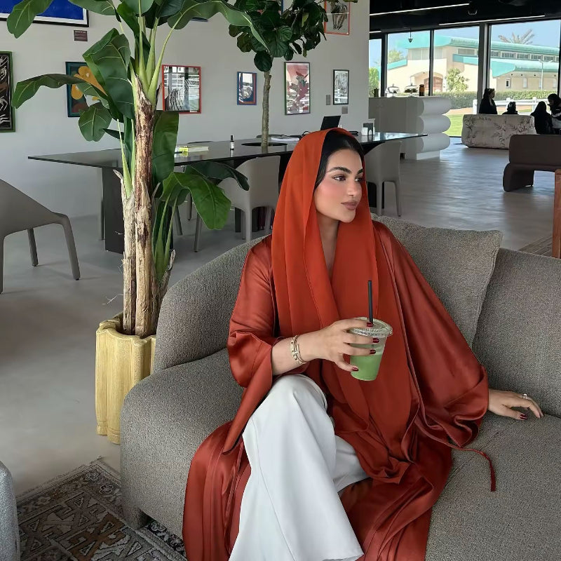 Side view of woman in rust orange ZAYNA abaya with hood, worn over a cream dress, accessorized with sunglasses.
