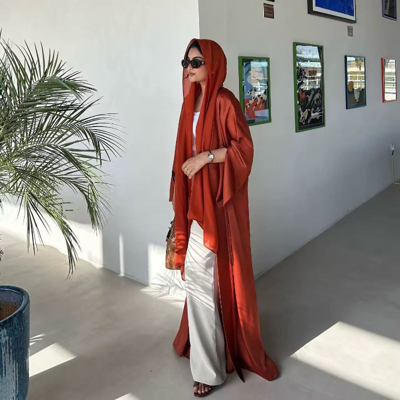 Side view of woman in rust orange ZAYNA abaya with hood, worn over a cream dress, accessorized with sunglasses.