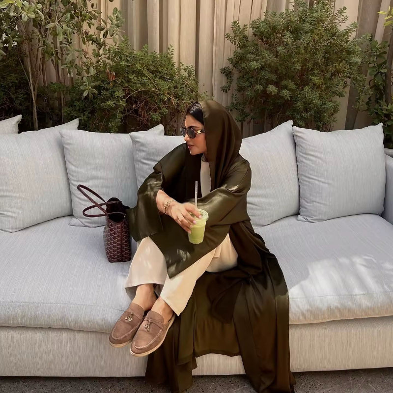 Woman wearing olive green ZAYNA satin abaya over a white outfit, holding a green drink, styled for summer.