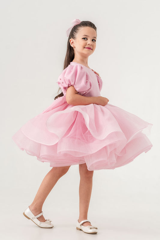 Luxury blush pink princess dress for toddler girls featuring elegant hand-sewn pearl embellishments on puff sleeves, a voluminous layered tulle skirt, and a dramatic oversized bow at the back. Perfect for birthdays, weddings, flower girl moments, and formal occasions. Soft 100% cotton lining for sensitive skin. Couture-inspired, handmade design for unforgettable childhood memories. Ideal for premium kids fashion, special occasion outfits, and baby girl party dresses. Elegant toddler outfit with invisible zi
