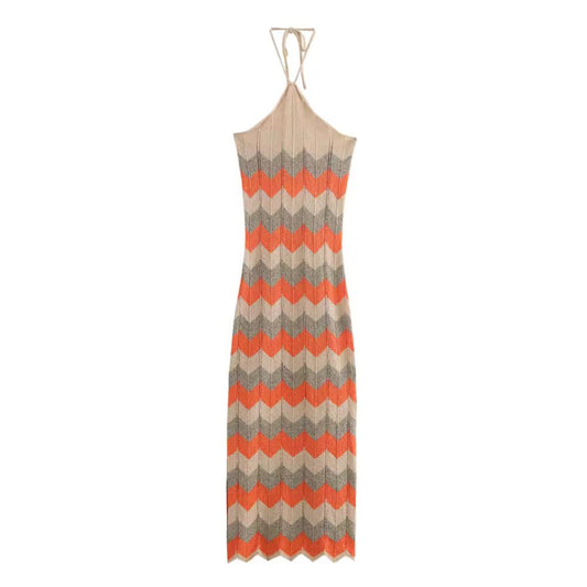 Woman wearing boho knit halterneck maxi dress in beige and orange zigzag pattern standing by a white wall in sunset light
