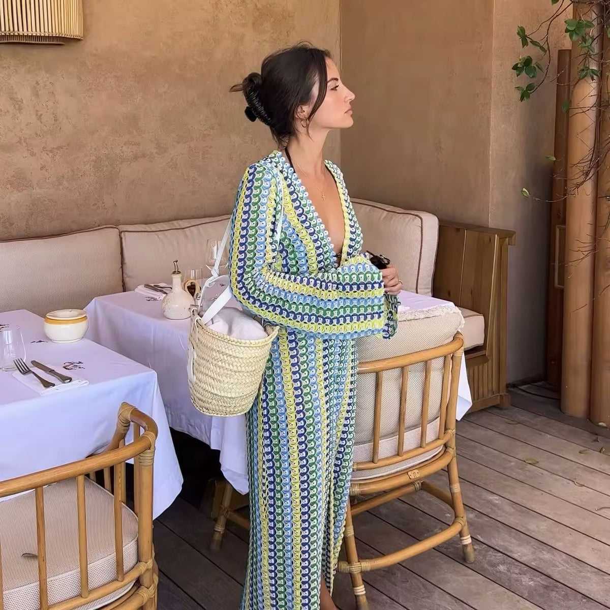 Woman wearing a colorful crochet knit maxi dress with vertical rainbow stripes, deep V-neckline, flared long sleeves, and high leg slit – standing confidently on a sunny beach, styled for summer vacations, resort wear, boho chic fashion, and Italian coastal vibes.