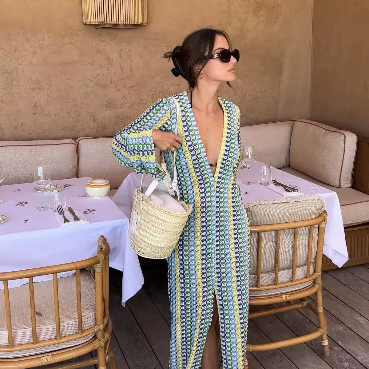 Woman wearing a colorful crochet knit maxi dress with vertical rainbow stripes, deep V-neckline, flared long sleeves, and high leg slit – standing confidently on a sunny beach, styled for summer vacations, resort wear, boho chic fashion, and Italian coastal vibes.