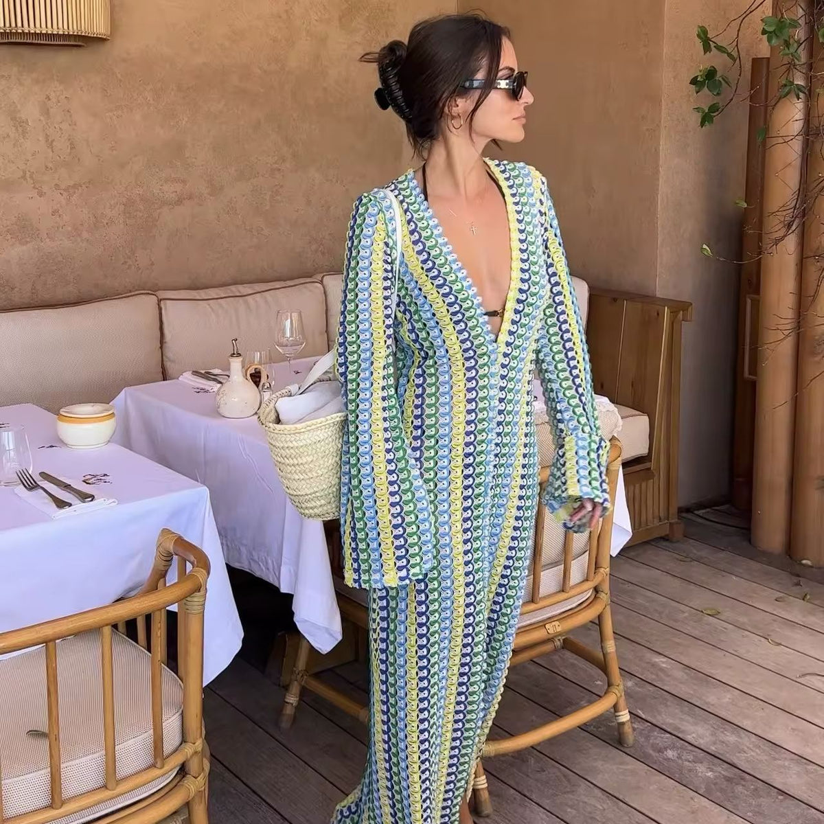 Woman wearing a colorful crochet knit maxi dress with vertical rainbow stripes, deep V-neckline, flared long sleeves, and high leg slit – standing confidently on a sunny beach, styled for summer vacations, resort wear, boho chic fashion, and Italian coastal vibes.