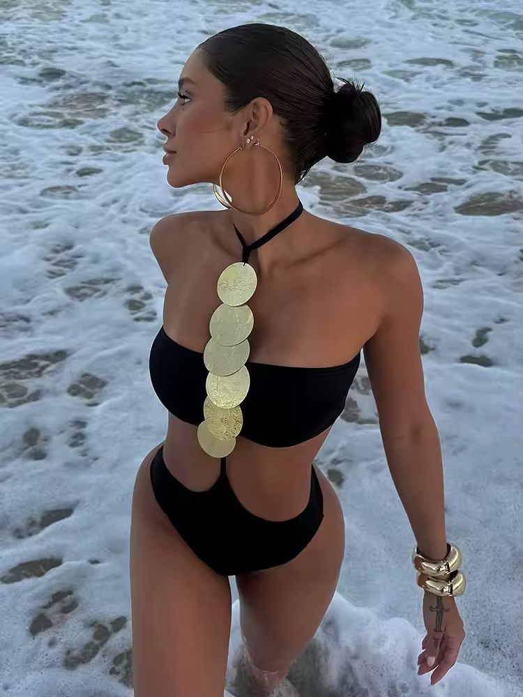 Woman wearing a sleek black one-piece swimsuit with gold statement discs and side cutouts, styled with hoop earrings and bold bangles on the beach during sunset – luxury swimwear look for summer, beach clubs, and resort style.