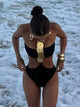Woman wearing a sleek black one-piece swimsuit with gold statement discs and side cutouts, styled with hoop earrings and bold bangles on the beach during sunset – luxury swimwear look for summer, beach clubs, and resort style.